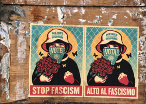 Posters of a woman in front of a chain-link fence holding roses, wearing a mask that says VOTE! and a hat that reads WE ARE HUMAN. A bold red bar at the bottom says STOP FASCISM. A Spanish version hang next to the English version.