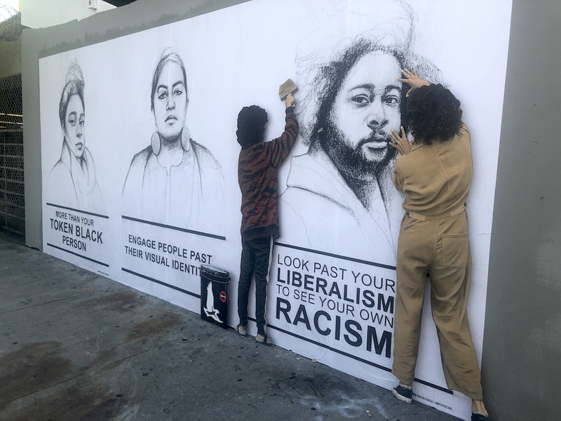 A mural with realist black-and-white sketches of people of color accompanied by messages like "Look past your liberalism to see your own racism." Freestanding representations of two artists appear to be placing the images onto the wall.