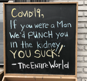 A handwritten chalkboard sign that reads "COVID-19, If you were a man we'd punch you in the kidney. YOU SUCK! - The Entire World"