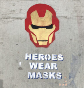 Stenciled sidewalk art of Iron Man's helmet with the text "HEROES WEAR MASKS" underneath