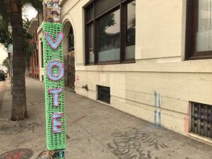 A street post wrapped in crochet that reads VOTE in vertical letters