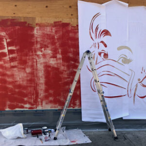 A plywood barrier with a large stencil attached to it and a ladder against it, as the artwork is being created.